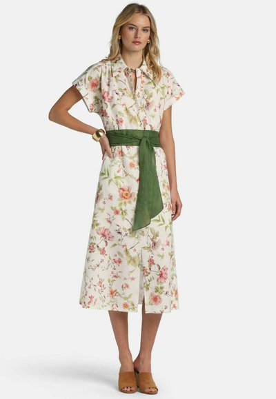 Mujer con vestido midi blanco con flores, cinturón de faja verde, mangas cortas, tacones abiertos marrones y pulsera y collar dorados.