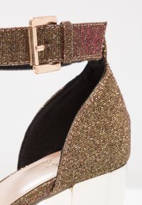 Gros plan d'une bride de cheville dorée pailletée avec une boucle métallique sur une chaussure à talon haut avec un talon blanc.