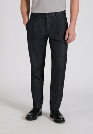 Mężczyzna ubrany w ciemnoszare plisowane spodnie, czarne skórzane mokasyny i jasnoszarą koszulę wpuszczoną do spodni, stojący na jednolitym tle.