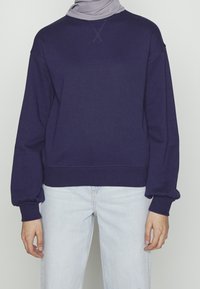 Osoba v tmavě modré mikině s dlouhým rukávem a světle modrých džínách, s světle šedým šátkem kolem krku.