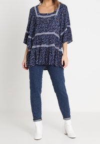 Plynulá námořnická modrá blůza s květinovým potiskem, krajkovými detaily a čtvercovým výstřihem. Spárována s modrými džíny a bílými kotníkovými botami.