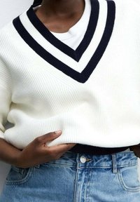 Personne portant un pull en maille blanc avec deux rayures noires sur le col en V, par-dessus une chemise blanche, rentrée dans un jean en denim bleu.
