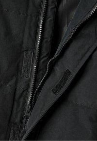 Veste noire isolée avec un tissu texturé, fermeture éclair visible et attache Velcro. La doublure intérieure est lisse et sombre.