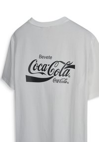 Witte T-shirt met zwarte opdruk "Bevete Coca-Cola" en het klassieke Coca-Cola-logo op de achterkant.