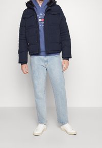 Marinblå puffjacka med flera framfickor, huva och ribbade cuffs, bärs över en blå hoodie och kombineras med ljusa denimjeans och vita sneakers.