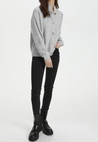 Graue Strickjacke mit Knopfleiste und Stehkragen, aus weichem Strickmaterial, getragen über schwarzen Skinny-Jeans und klobigen schwarzen Stiefeln.