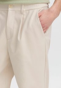 Man wearing beige pants with a hand tucked into the front pocket, visible button and waistband detail.