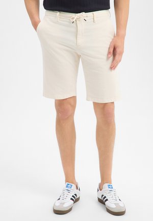 Mann trägt beige Shorts mit Kordelzug, schwarzes Oberteil und weiße Adidas-Sneaker, steht mit Händen in den Taschen auf weißem Hintergrund.