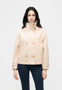 Γυναίκα που φοράει μπεζ διπλής σειράς μάλλινο παλτό με ψηλό γιακά και ξύλινα κουμπιά, συνδυασμένο με σκούρο εφαρμοστό παντελόνι.