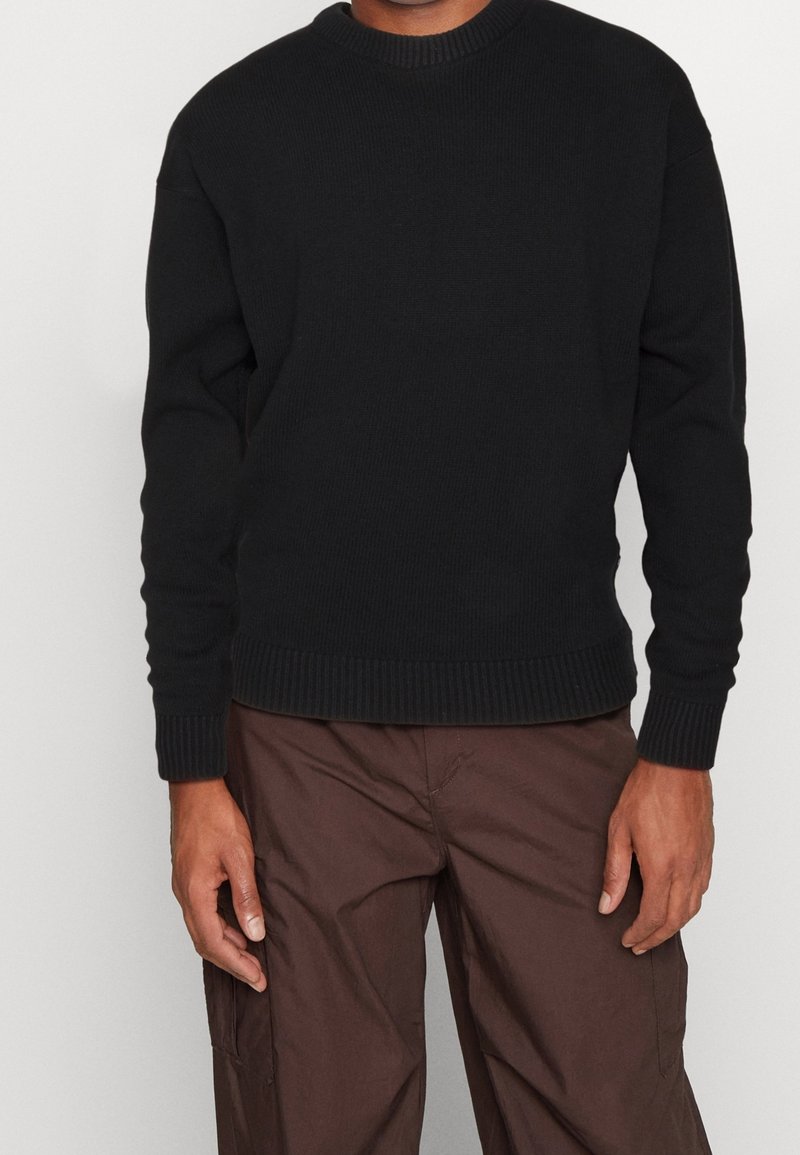 Pull noir en maille avec col rond, poignets et ourlet côtelés, présentant un tissu texturé. Porté sur un pantalon cargo marron.