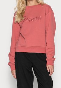 Žena v dlouhém růžovém svetru s dlouhým rukávem a výšivkou "Guess" na hrudi, spolu s volnými černými kalhotami.