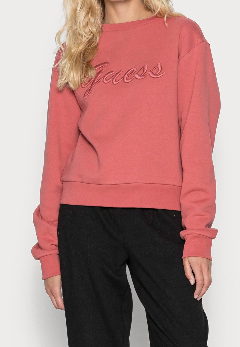 Žena v dlouhém růžovém svetru s dlouhým rukávem a výšivkou "Guess" na hrudi, spolu s volnými černými kalhotami.