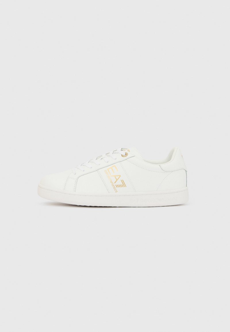 Fehér sportcipő sima szintetikus felsőrésszel, arany logóval az oldalon, lapos fűzőkkel és fehér gumitalppal. Finom varrási részlet.