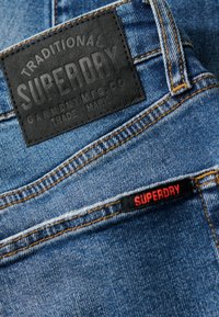 Vaqueros de mezclilla azul con un parche de cuero negro con logo en relieve y una etiqueta roja. Se aprecian costuras en contraste y detalles de bolsillos en ángulo.