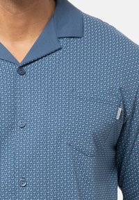 Lyhythihainen sininen paita, jossa on vaaleansininen geometrinen kuvio, teräväkauluksella, nappikiinnityksellä ja rinnassa taskulla.