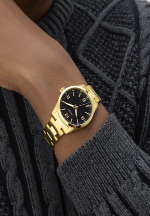 Goldfarbene Armbanduhr mit schwarzem Zifferblatt, numerischen Stundenmarkierungen und einem Metall-Armband, getragen an einer Hand gegen einen grau gestrickten Pullover.