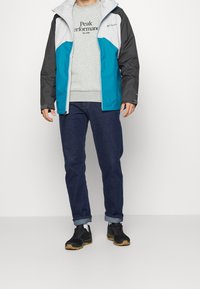Water-resistant jacket with a gray, turquoise, and white color block design. Features a hood and zip front, paired with blue jeans and black sneakers.