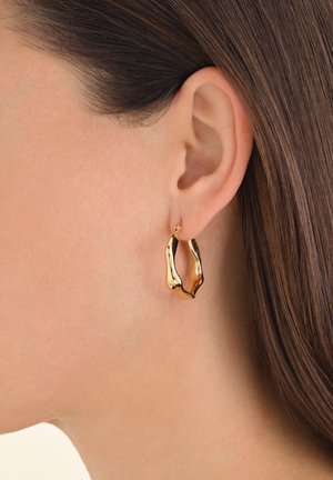 Boucles d'oreilles en or, abstraites, avec un design ondulé, présentant des surfaces brillantes et polies qui reflètent la lumière. Petits fermoirs à clous visibles.