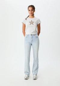 Siva majica s grafiko rjave zvezde, v kombinaciji s svetlo modrimi širokimi džinsom. Džins so oblikovani v klasičnem pet-pocket dizajnu z gumbom.