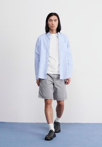 Camisa de cuadros azul claro sobre una camiseta blanca, combinada con pantalones cortos grises y zapatos negros texturizados. Estilo casual y ajuste relajado.