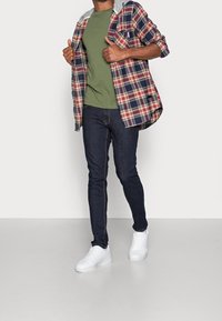 Model v zeleném tričku, rozepnuté červené a modré kostkované košili se šedou kapucí, tmavých džínách a bílých teniskách.