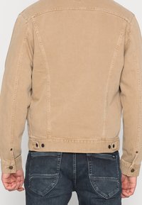 Vaaleanruskea farkkutakki, jossa on muotoiltu siluetti, näkyvät ompeluyksityiskohdat ja napilliset hihansuut, yhdistettynä tummansinsisiin farkkuihin.