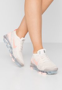 Scarpe da ginnastica in mesh rosa chiaro con tomaia in maglia, suola trasparente con ammortizzazione ad aria e dettagli riflettenti argentati vicino ai lacci.