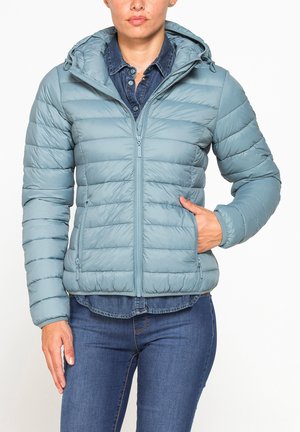 Persona che indossa una giacca imbottita azzurro chiaro sopra una camicia di jeans blu scuro e jeans, con una mano nella tasca della giacca e l'altra di lato.