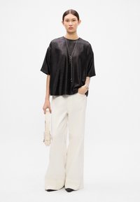 Černý metalický oversized top s krátkým rukávem, skombinovaný s bílými širokými kalhotami s vysokým pasem. Doplňky zahrnují stříbrný náhrdelník a krémovou kabelku.