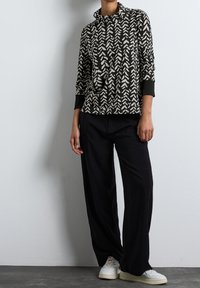 Černý a béžový vzorovaný top s vysokým límcem a dlouhými rukávy; v kombinaci se volnými černými kalhotami a bílými teniskami na texturované šedé podlaze.