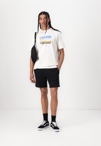 T-shirt crème avec un imprimé graphique coloré, assortie à un short en jean noir. Un sac messager noir, des chaussettes noires et des baskets noir et blanc complètent le look.