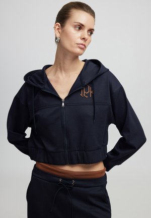 Marineblauwe cropped hoodie van zachte stof met een rits aan de voorkant, voorzien van een geborduurd logo en trekkoorden op de kap. Gepaard met bijpassende broek.