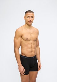 Μυώδης άντρας που φοράει μαύρα εσώρουχα Tommy Hilfiger boxer, στέκεται μπροστά σε ένα απλό ανοιχτό γκρι φόντο, στραμμένος ελαφρώς προς τα αριστερά.