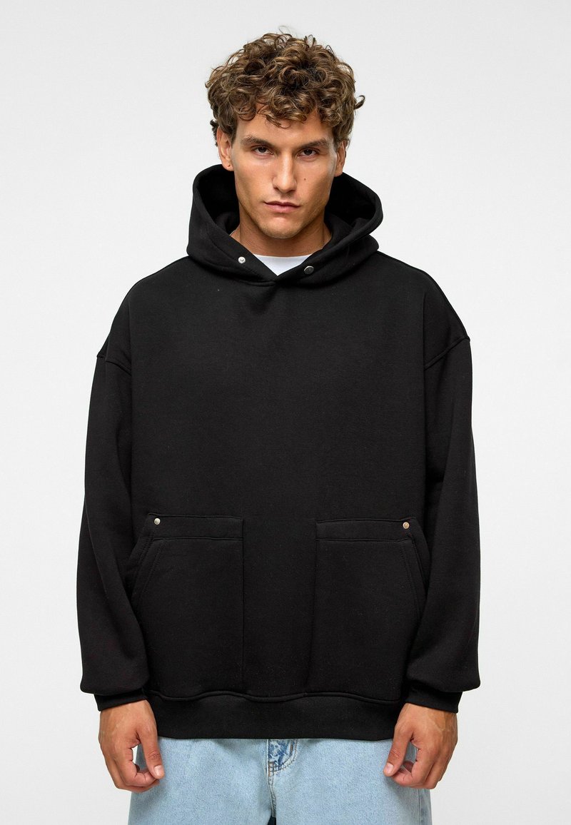 Zwarte capuchontrui van zacht materiaal, met een losse pasvorm, twee voorzakken en decoratieve drukknoopjes op de zakken.