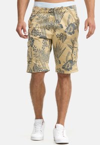 Shorts cargo beige avec un motif floral gris foncé, dotés de plusieurs poches et d'une taille plate avec le nom de la marque en bleu clair.