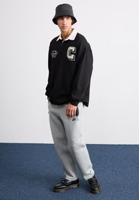 Polo negru cu mânecă lungă și guler alb, detaliu grafic; pantaloni de trening gri cu logo; pantofi negri; purtat cu o pălărie bucket neagră.