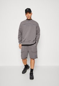 Grijze sweatshirt met geborduurd logo, gecombineerd met bijpassende grijze shorts met voorzakken. Een zwarte pet en schoenen maken de outfit af.