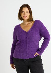Cárdigan morado esponjoso con escote en V, mangas largas y botones en la parte delantera; usado sobre jeans oscuros. Textura suave y corte relajado.
