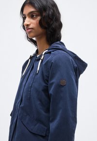 Ναυτικό σακάκι με κουκούλα από λεία ύφανση, με φερμουάρ, κορδόνια, ένα έμπλαστρο στο α ριστερό μπράτσο και πλαϊνές τσέπες.