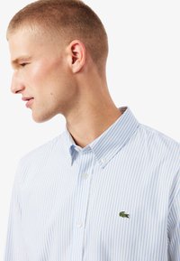 Camisa de botones a rayas azul claro con un collar clásico y un pequeño logo de cocodrilo bordado en el lado izquierdo del pecho. Textura de tela suave.