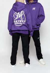 Lila Kapuzenpulli mit weißem Text "MULTIPLY APPAREL 15" auf dem Rücken, kombiniert mit schwarzen Hosen. Der Kapuzenpulli verfügt über eine Kängurutasche und eine Kapuze.