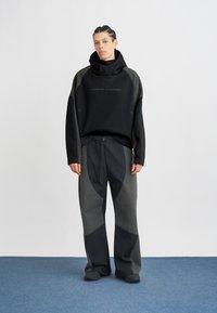 Fiatal személy áll, fekete és sötétszürke Nike Tech kapucnis pulóvert és azzal megegyező nadrágot visel, kék szőnyegen, egyszínű fehér háttér előtt.