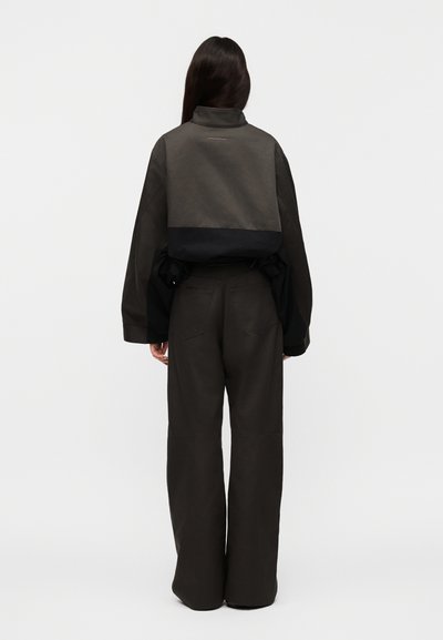 Άτομο με μακριά σκούρα μαλλιά που φοράει ένα διχρωμο σκούρο κοντό μπουφάν και φαρδιά σκούρα παντελόνια, γυρισμένο πλάτη σε ένα απλό ανοιχτόχρωμο φόντο.