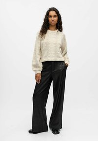 Cremefarbener, kabelstrickgestrickter Pullover mit Puffärmeln, kombiniert mit hochtaillierten, glitzernden schwarzen Hosen und schwarzen Schuhen, vor einem neutralen Hintergrund.