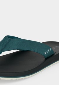 Μαύρη σαγιονάρα με τιρκουάζ υφασμάτινο λουράκι με το κείμενο «REEF», υφασμένο πέλμα και λευκή αυλακωτή σόλα.