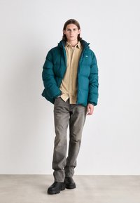 Giacca puffer verde acqua con cappuccio, dotata di zip e polsini elastici, abbinata a una camicia beige e jeans grigi, completata da stivali neri.