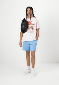 Lyserød grafisk T-shirt med tegneserie design, lyseblå shorts, sort crossover taske, hvide sneakers og grå strømper. Enkel, afslappet outfit.