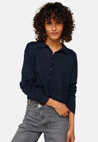 Chemise bleu marine à boutons avec un col, manches longues et une coupe courte. Tissu doux au toucher lisse, associée à un jean gris.
