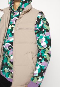 Chaleco acolchado beige con cuello alto, que incluye un bolsillo con cremallera. Debajo, una sudadera con capucha de camuflaje multicolor con puños morados.