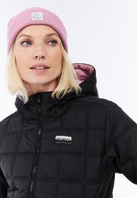 Giubbotto imbottito nero trapuntato con cappuccio e chiusura a zip, con una toppa bianca con logo. Abbinato a un berretto in maglia rosa chiaro.